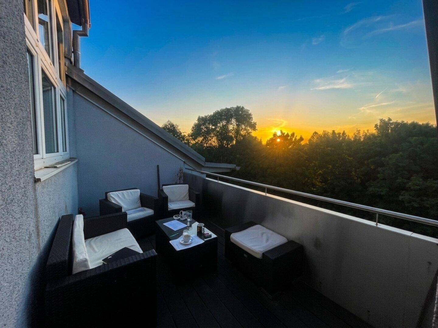 Gemütlicher Balkon mit Sitzgelegenheiten und einem wunderschönen Sonnenuntergang über den Bäumen.