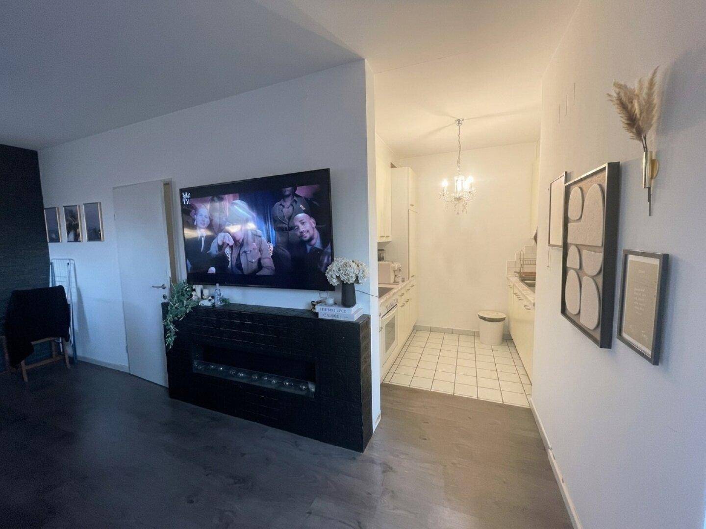 Wohnbereich mit großem Fernseher über einer dunklen Konsole, angrenzend an eine kleine Küche.