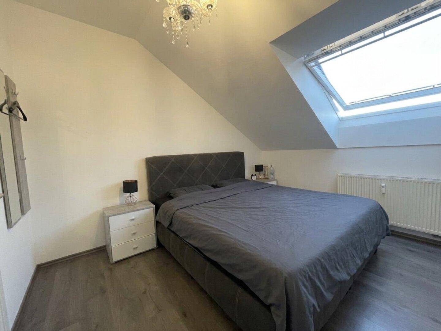 Schlafzimmer mit grauem Bett, Nachttischen und einem großen Dachfenster in der Dachschräge.