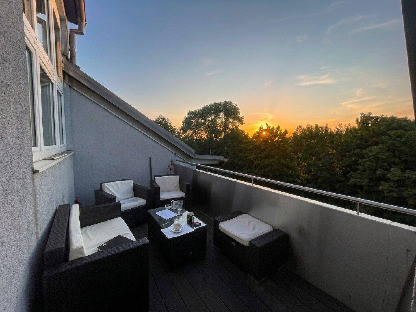 Gemütlicher Balkon mit Sitzgelegenheiten und einem wunderschönen Sonnenuntergang über den Bäumen.