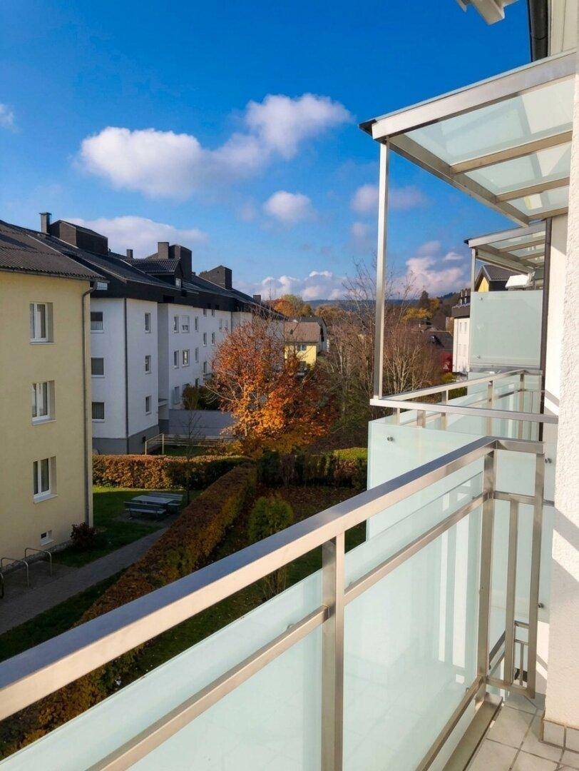 Balkon mit Glasgeländer und Blick auf Wohngebäude sowie herbstliche Bäume.
