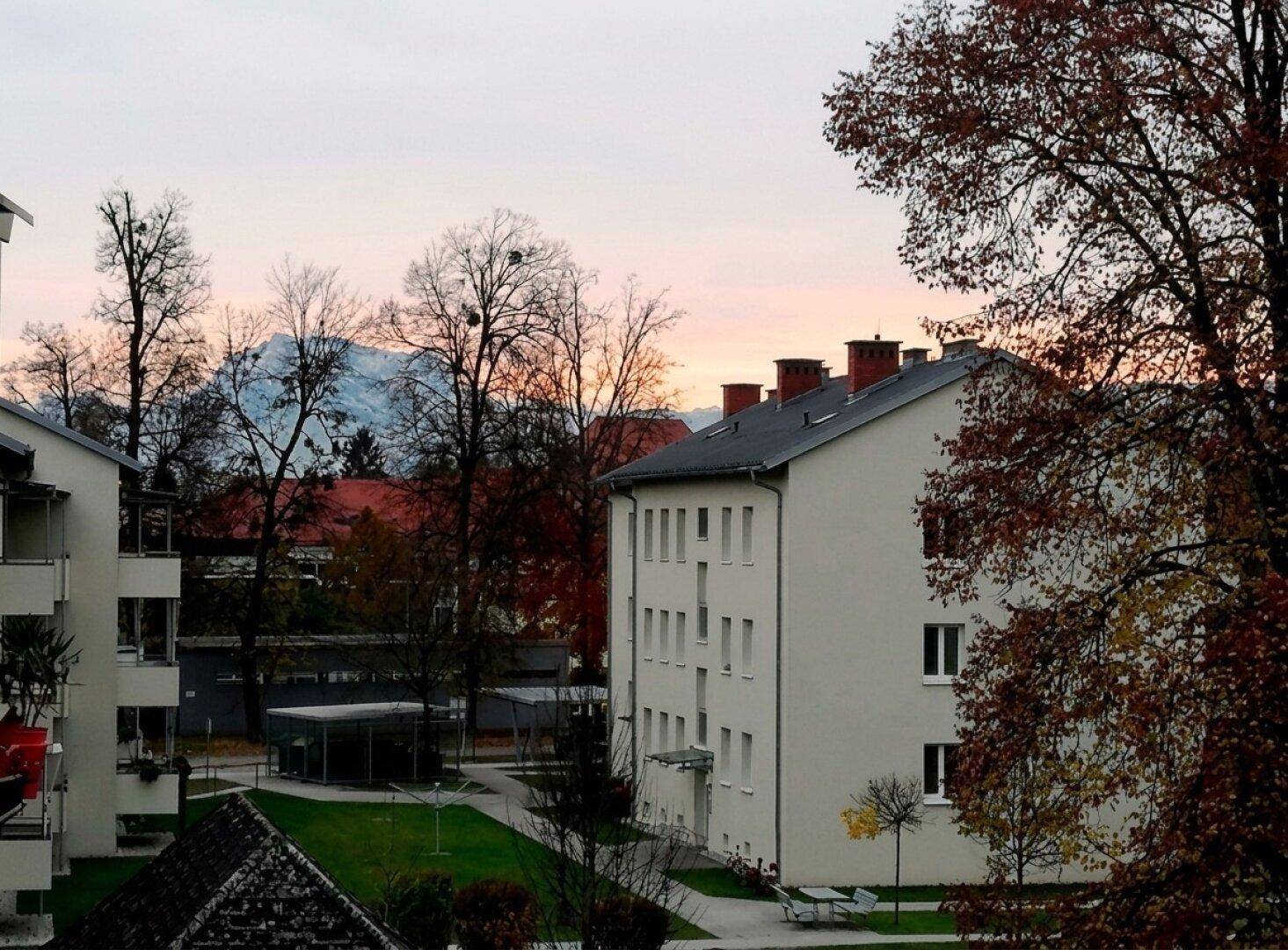 Abendlicher Blick auf Wohngebäude, Grünflächen und schneebedeckte Berge in der Ferne.