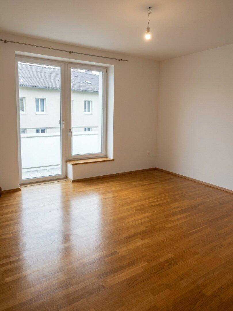 Leerer Raum mit hellem Holzboden und großer Balkontür, die viel Licht hereinlässt.