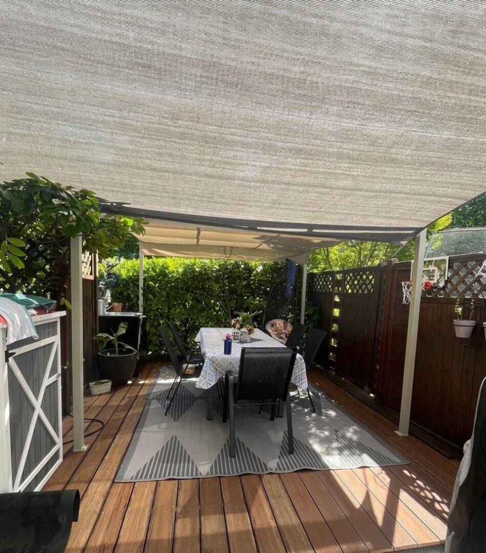 Gemütliche überdachte Terrasse mit Esstisch und Holzdeck, umgeben von grüner Hecke.