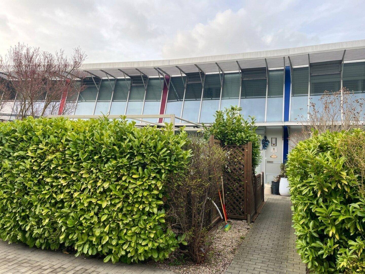 Moderne Reihenhausfassade mit großen Fenstern und gepflegter grüner Hecke.