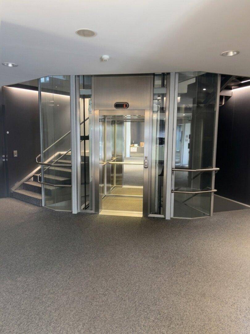 Moderner Eingangsbereich mit einem gläsernen Aufzug und einer eleganten Treppe, die zu den oberen Etagen führt.