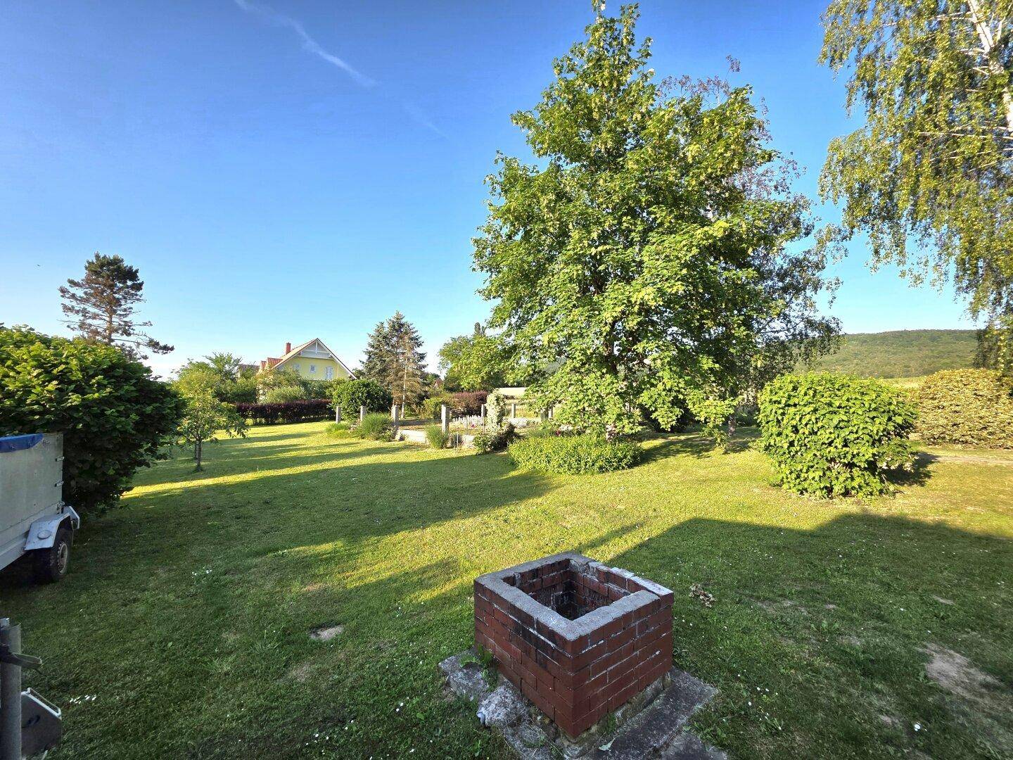 Weitläufiger grüner Garten mit großen Bäumen und einem gemauerten Grillplatz unter strahlendem Himmel.