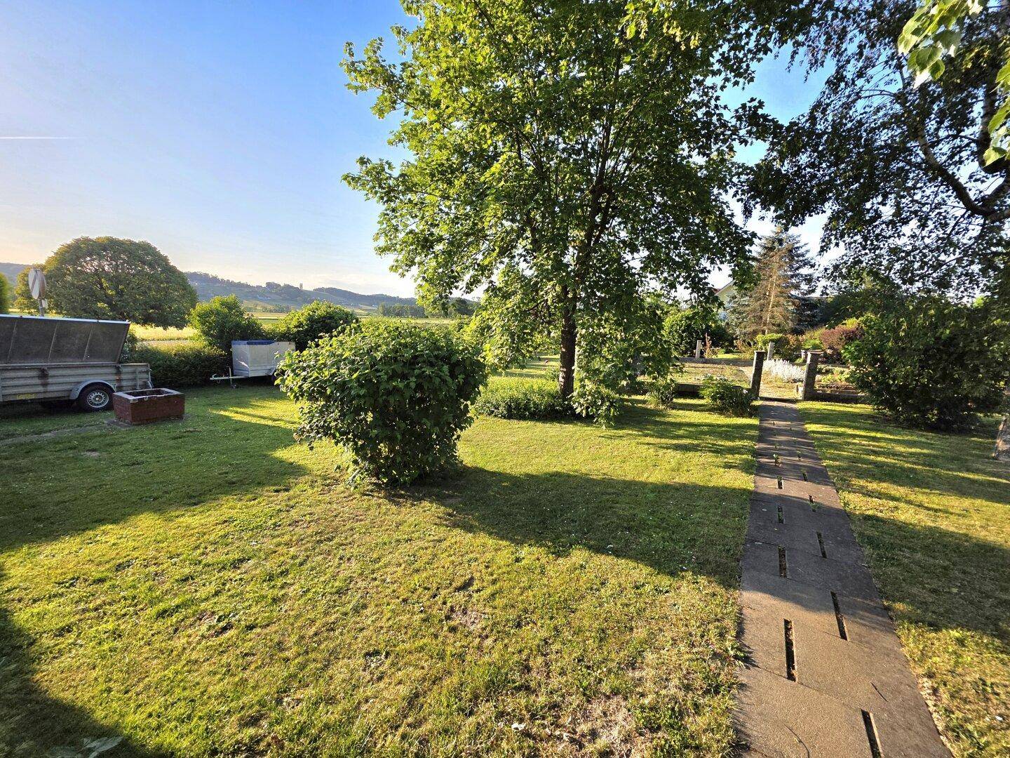 Großer, sonniger Garten mit Rasenfläche, Bäumen und einem Steinweg, der durch das Grün führt.
