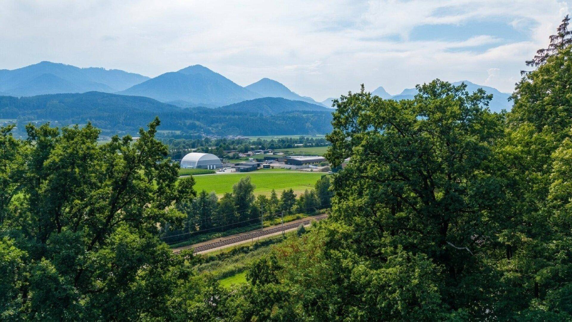 Weitblick über eine grüne Landschaft mit Bergen, Feldern und einer Bahnlinie im Vordergrund.