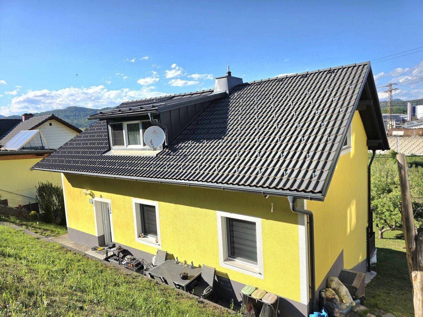 Gelbes Einfamilienhaus mit dunklem Ziegeldach und gepflegter Fassade unter blauem Himmel.