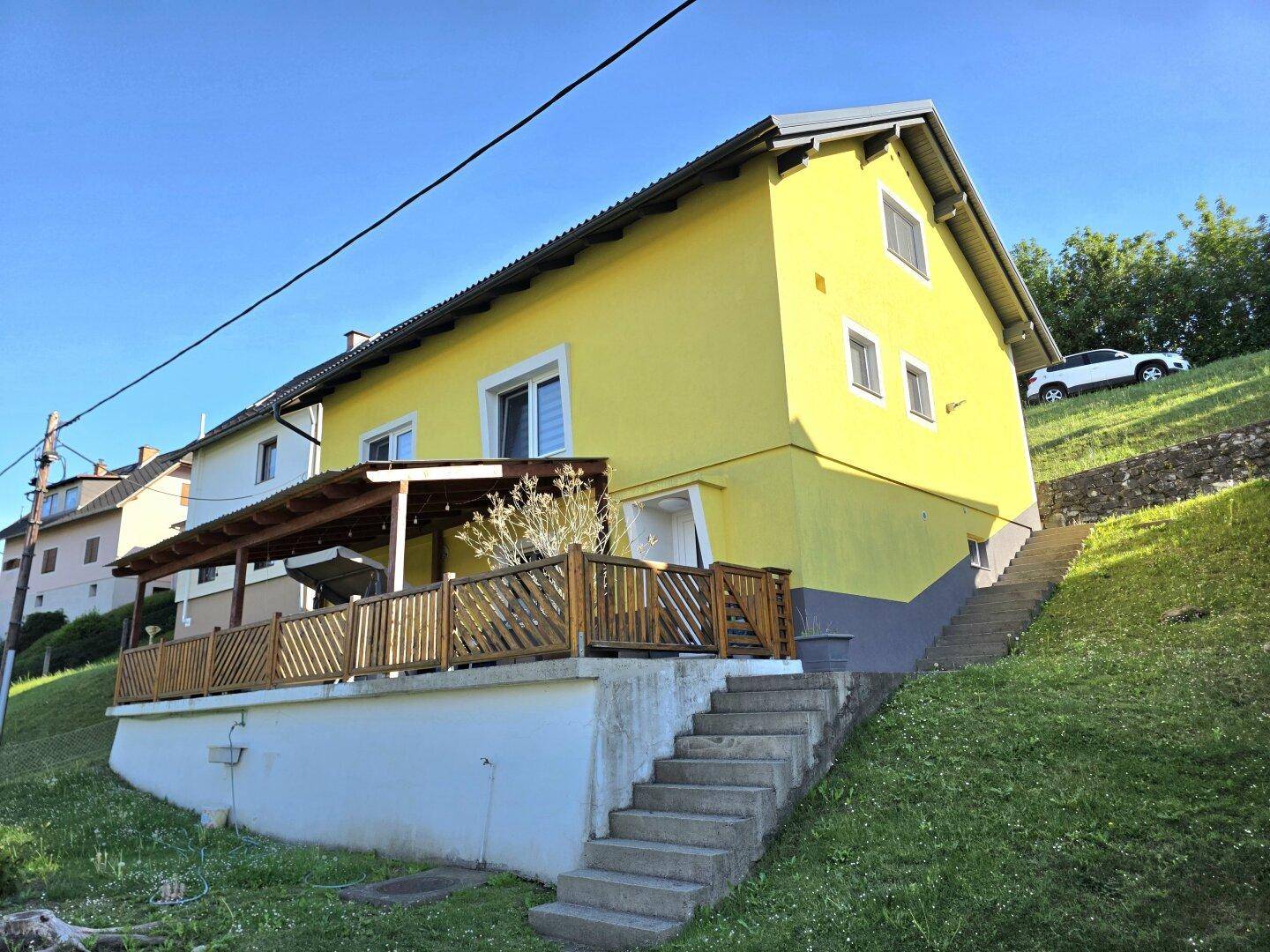 Gelbes Haus mit überdachter Holzterrasse und Treppe, umgeben von grüner Landschaft.