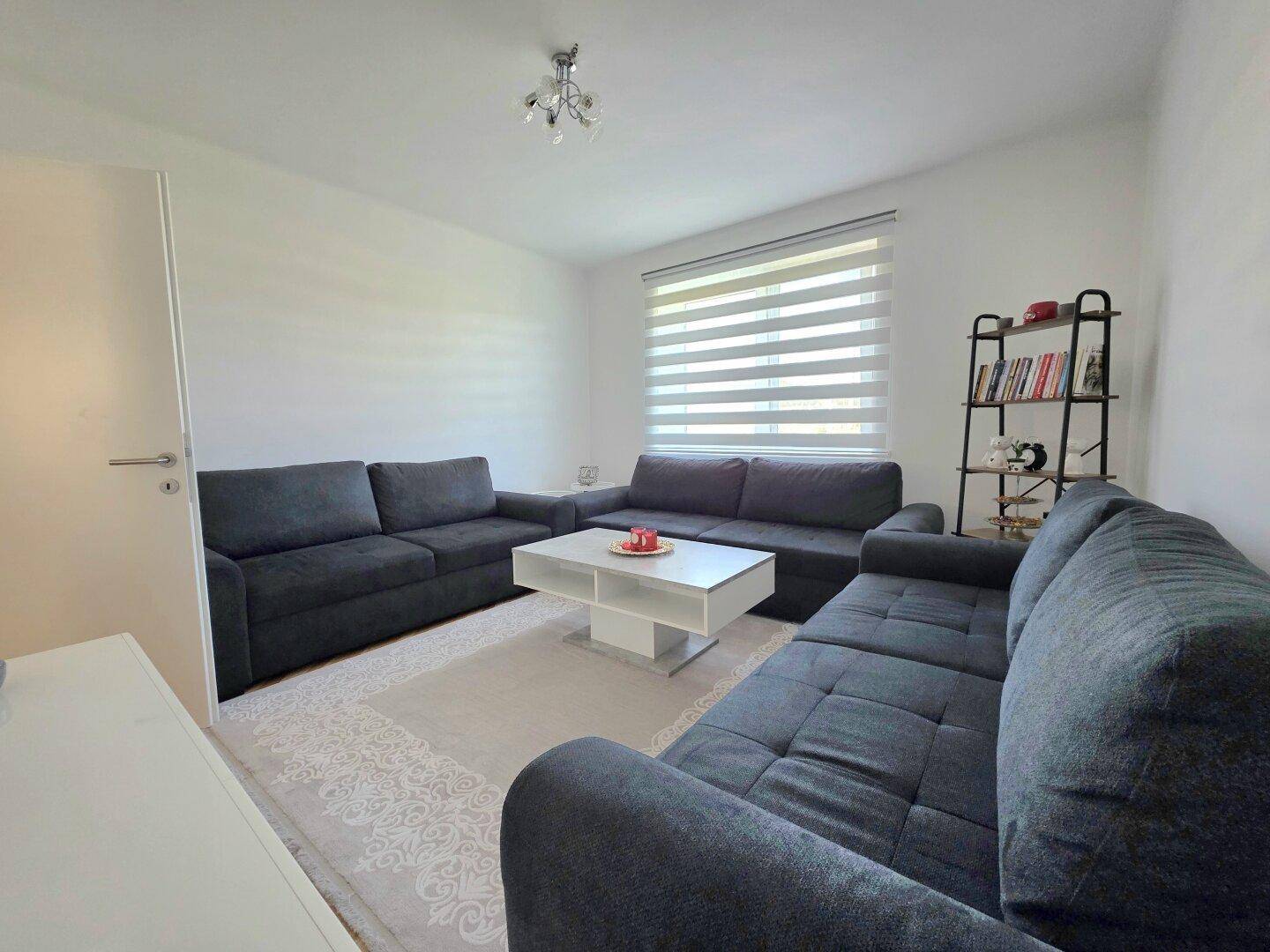 Gemütliches Wohnzimmer mit großen Sofas, hellem Teppich und Fenster mit Jalousien.