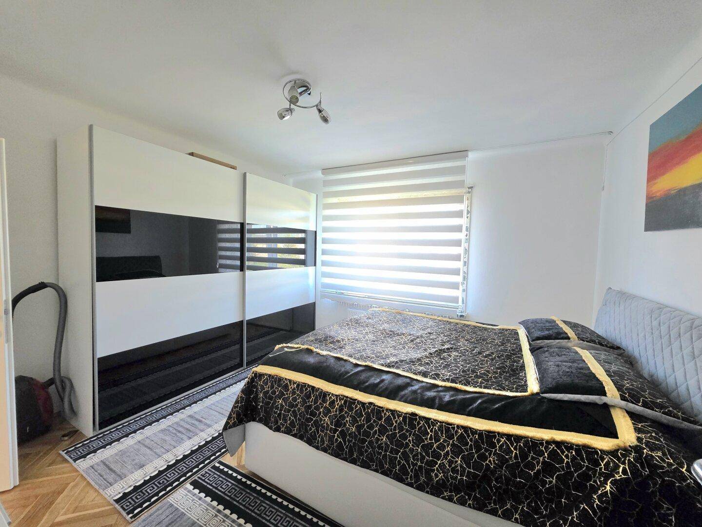 Schlafzimmer mit modernem Kleiderschrank, Doppelbett und hellem Fenster mit Jalousien.