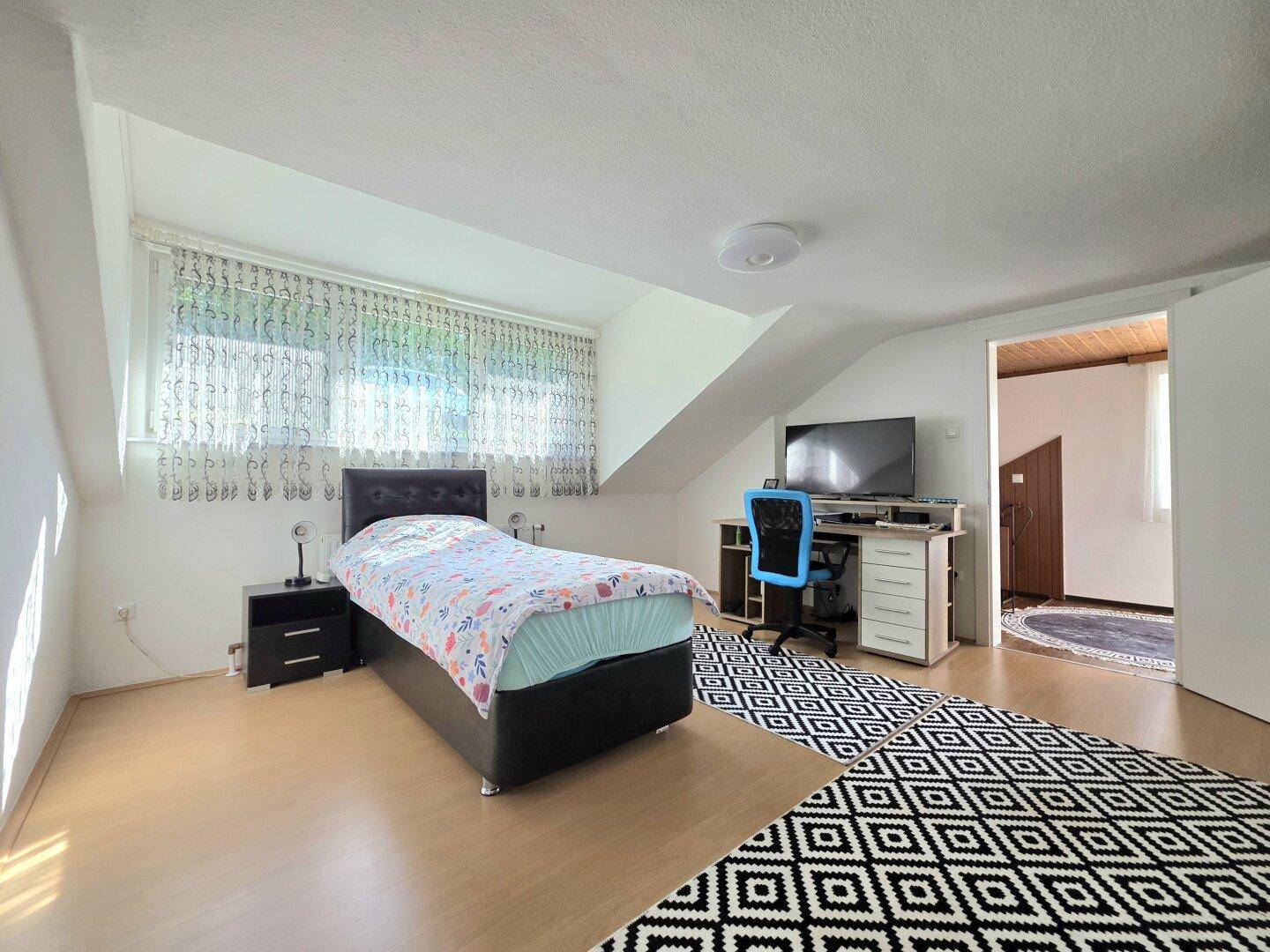 Helles Dachgeschoss-Schlafzimmer mit Einzelbett, Schreibtisch und gemustertem Teppich.