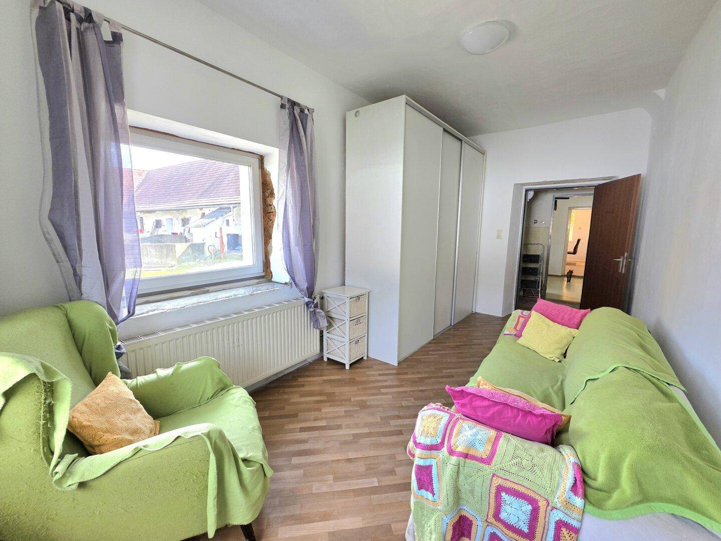 Helles Zimmer mit grünem Sofa, Kleiderschrank und Laminatboden, ideal als Wohn- oder Gästezimmer.