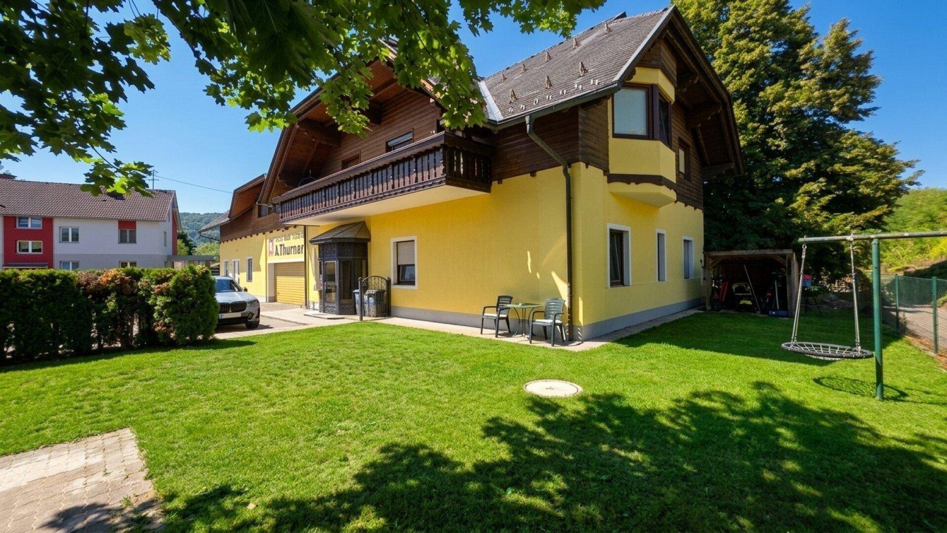 Gelbes Gebäude mit Holzbalkon, gepflegtem Rasen und Kinderschaukel im Vorgarten.