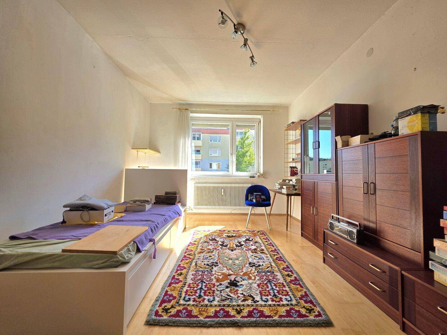 Helles Schlafzimmer mit Einzelbett, Holzmöbeln und farbenfrohem Teppich.