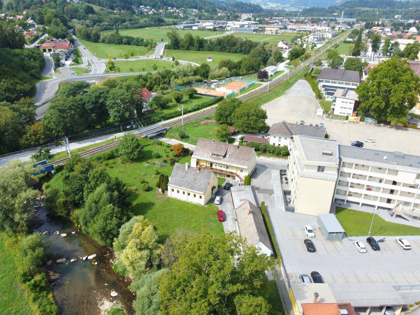 Luftaufnahme einer ländlichen Umgebung mit Häusern, einem Fluss und Sportanlagen im Hintergrund.
