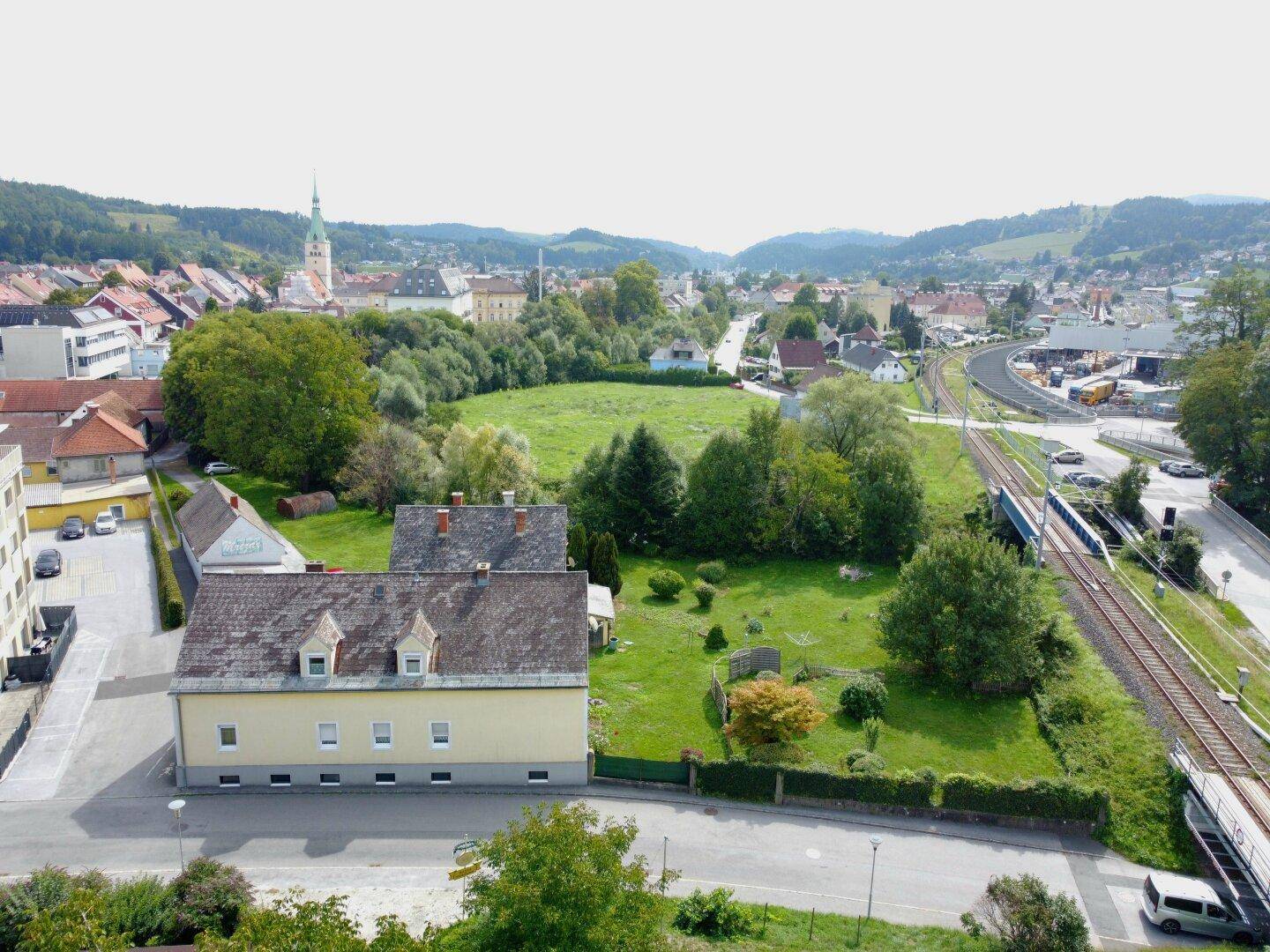 Weitläufige Luftaufnahme einer Stadt mit Wohngebäuden, viel Grün und einer Bahnlinie.