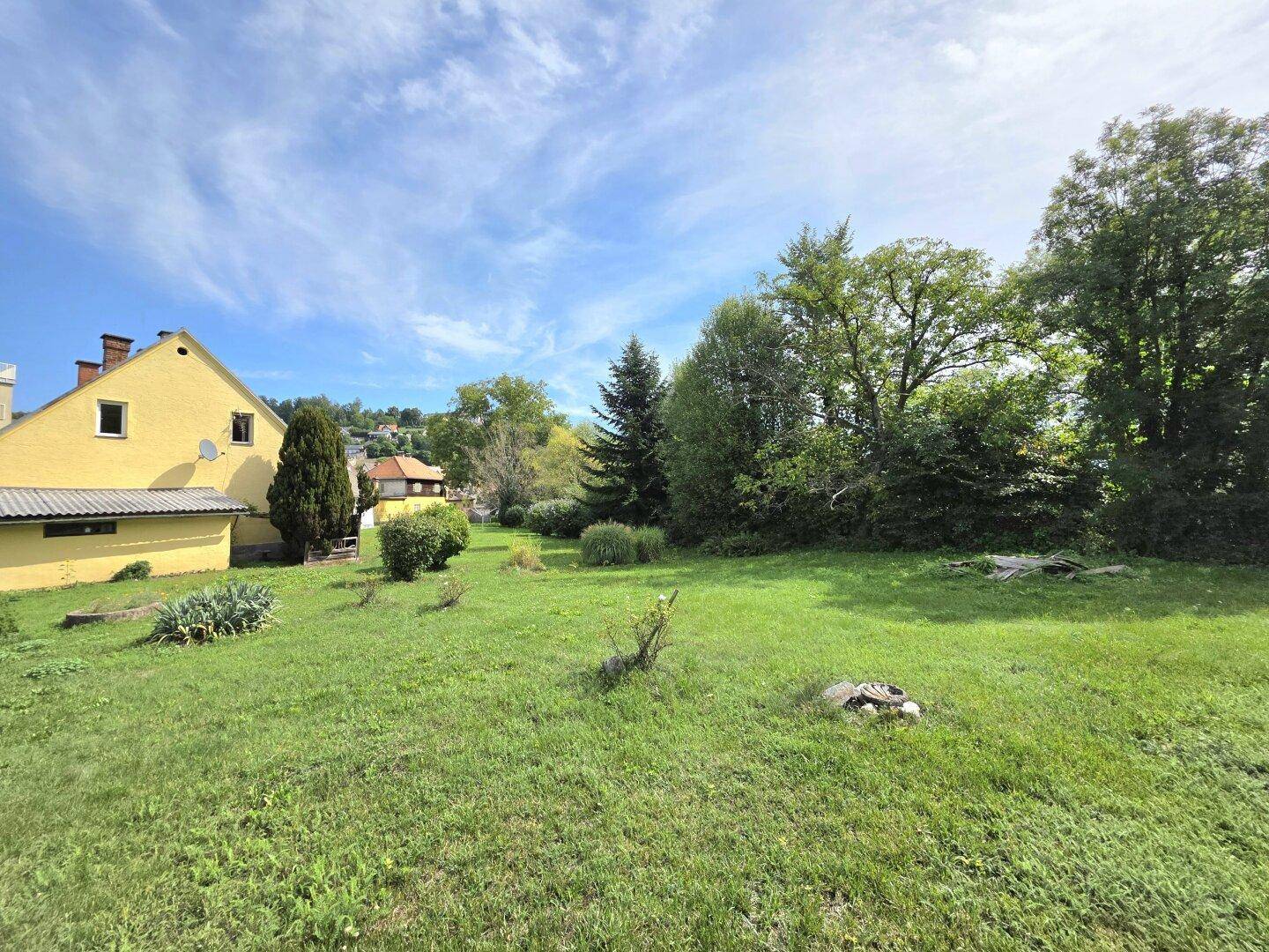 Weitläufiger Garten mit grüner Rasenfläche, verschiedenen Bäumen und einem gelben Gebäude im Hintergrund.