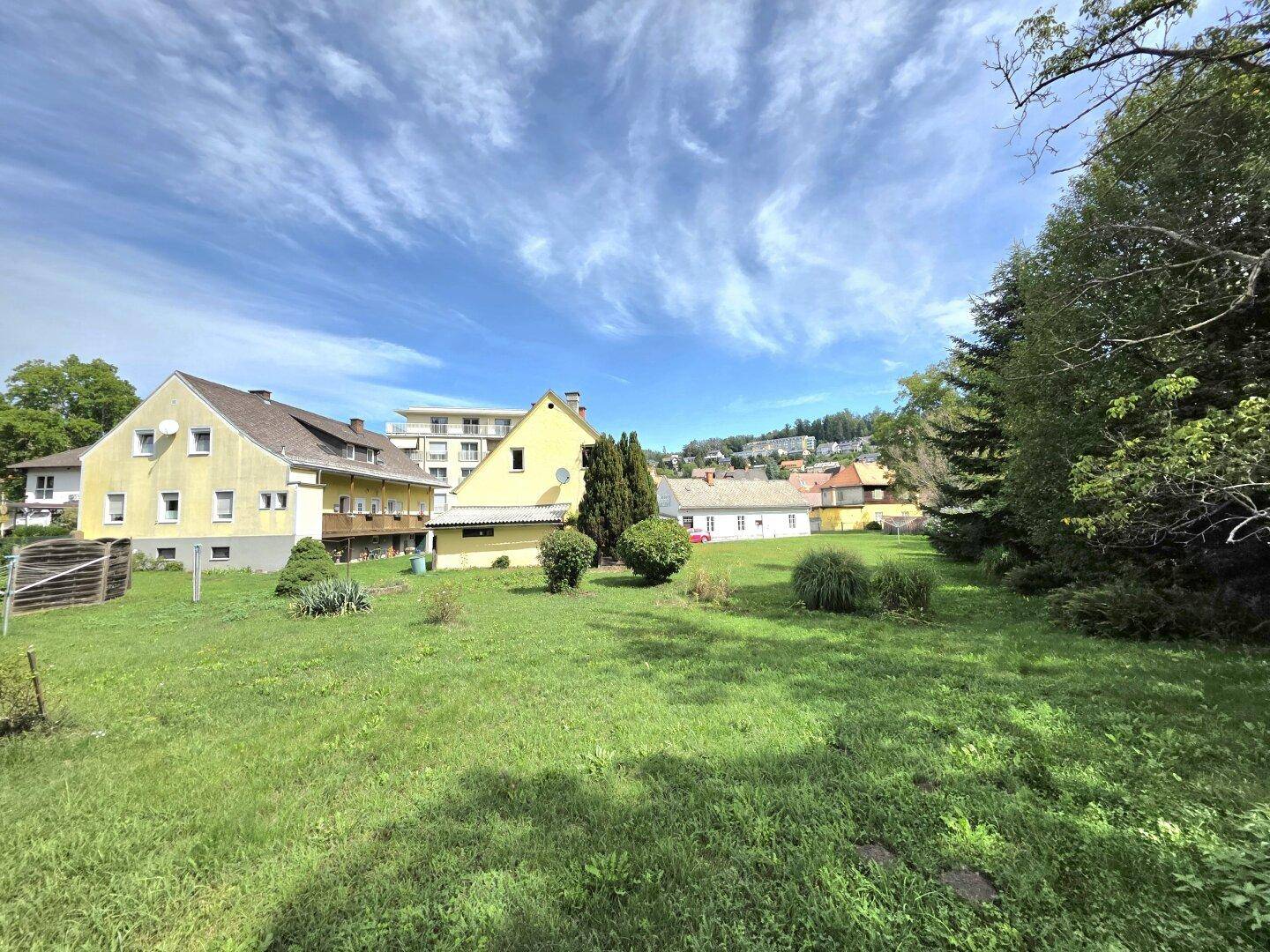 Sonniger Garten mit gepflegtem Rasen, hohen Bäumen und mehreren Gebäuden im Hintergrund.