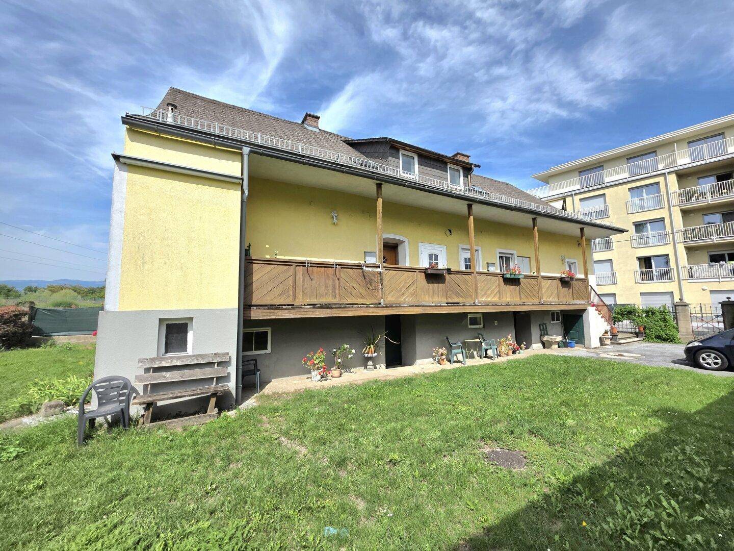 Rückseite eines Gebäudes mit einem langen Holzbalkon und einer kleinen Rasenfläche mit Sitzgelegenheiten.