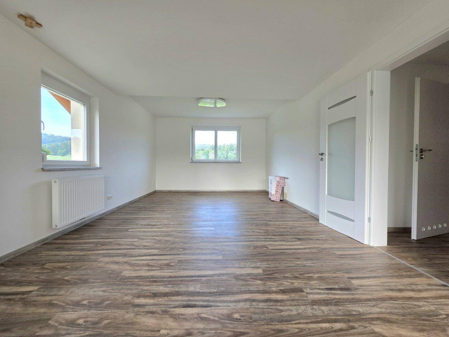 Geräumiger, unmöblierter Raum mit Laminatboden und Fenstern mit Blick ins Grüne.