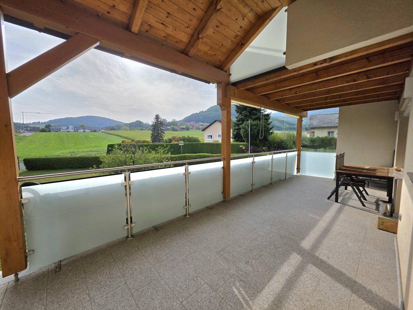 Geräumiger überdachter Balkon mit Glasgeländer und weitem Blick über die Landschaft.