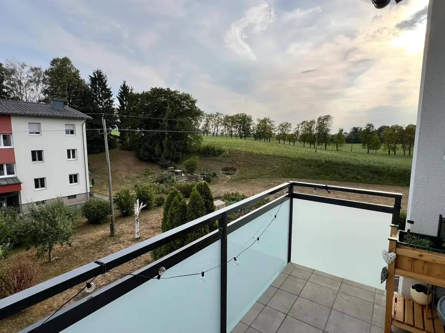 Balkon mit Glasbrüstung und Blick auf grüne Wiesen und Bäume unter bewölktem Himmel.