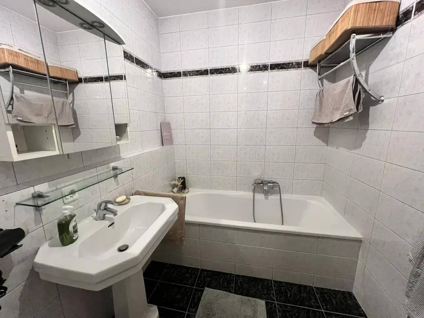 Helles Badezimmer mit Badewanne, Waschbecken und weißen Fliesen, akzentuiert durch eine schwarze Bordüre.