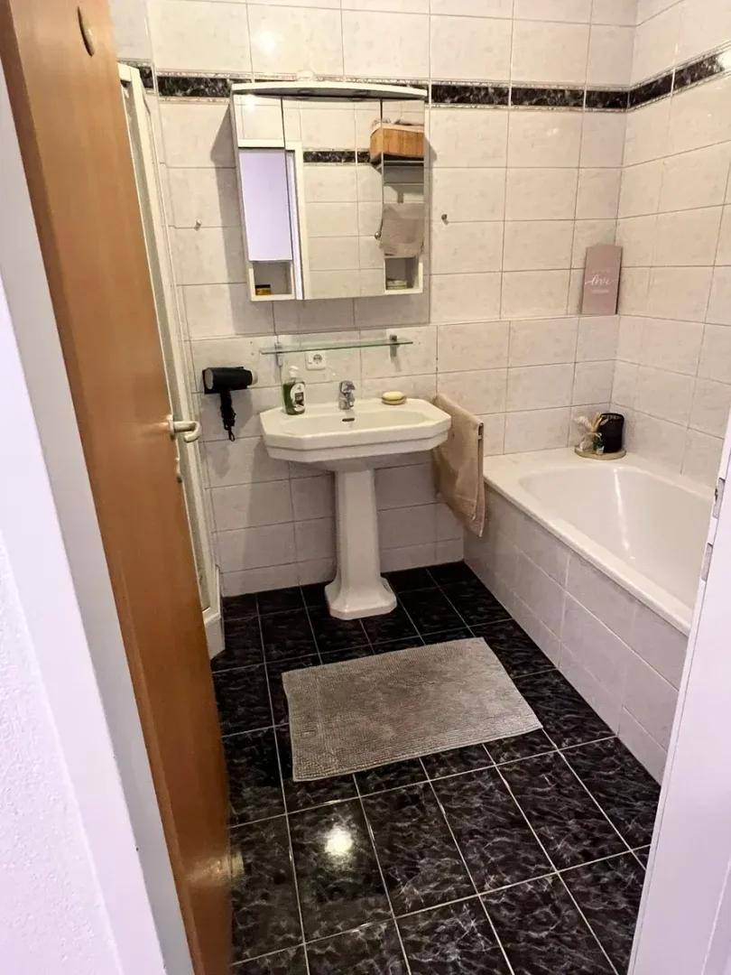 Badezimmer mit Badewanne, Waschbecken und Spiegel, ausgestattet mit weißen Fliesen und dunklem Boden.