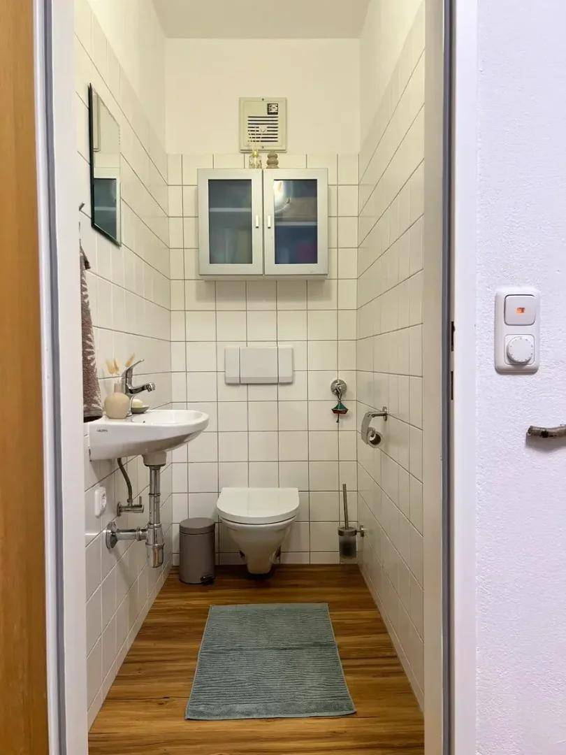 Kleines, hell gefliestes WC mit Waschbecken, Toilette und Holzoptikboden.