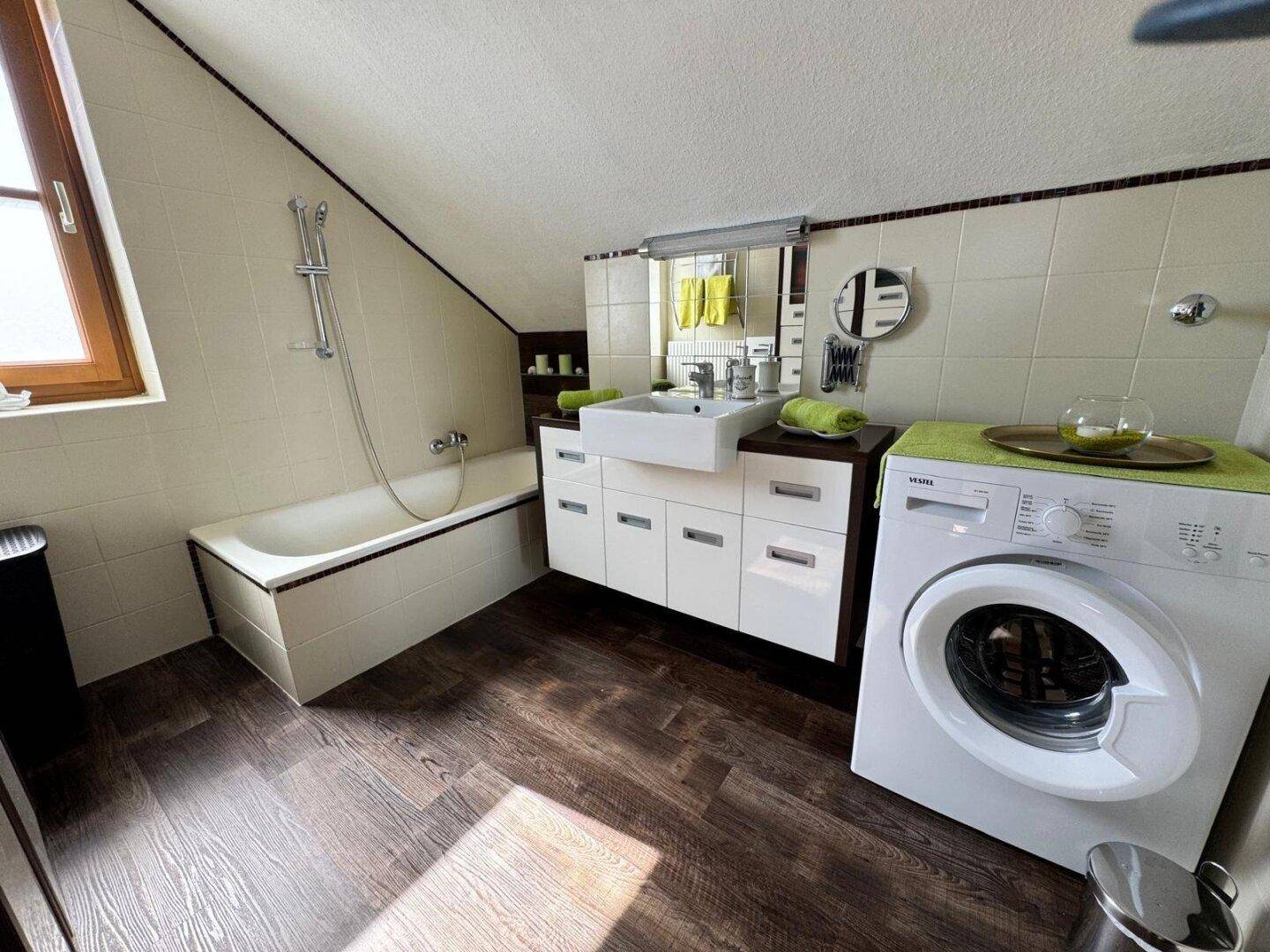 Helles Badezimmer mit Badewanne, Waschbecken und Waschmaschine unter Dachschräge.