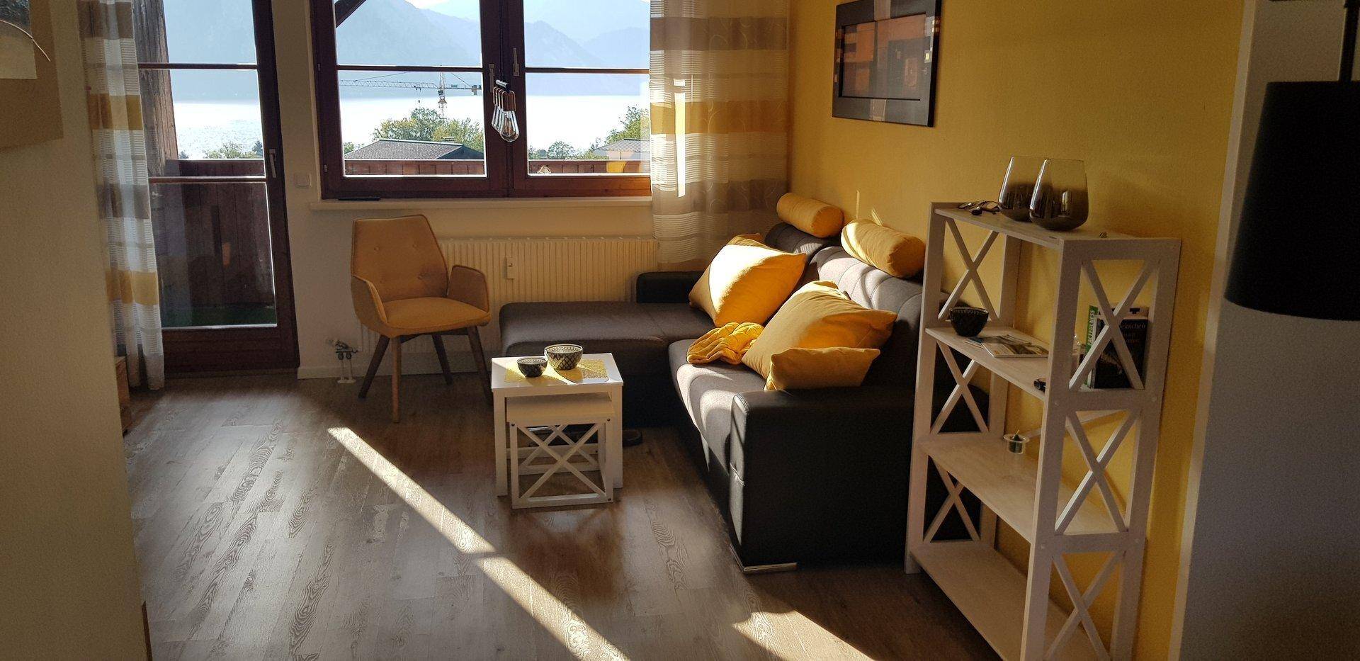 Gemütliches Wohnzimmer mit Seeblick, gelben Kissen und viel natürlichem Licht.