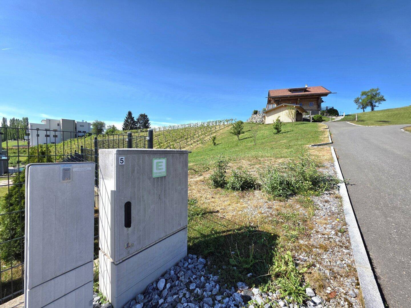 Blick auf eine grüne Landschaft mit Weinberg, einem Holzhaus und Versorgungsboxen im Vordergrund.