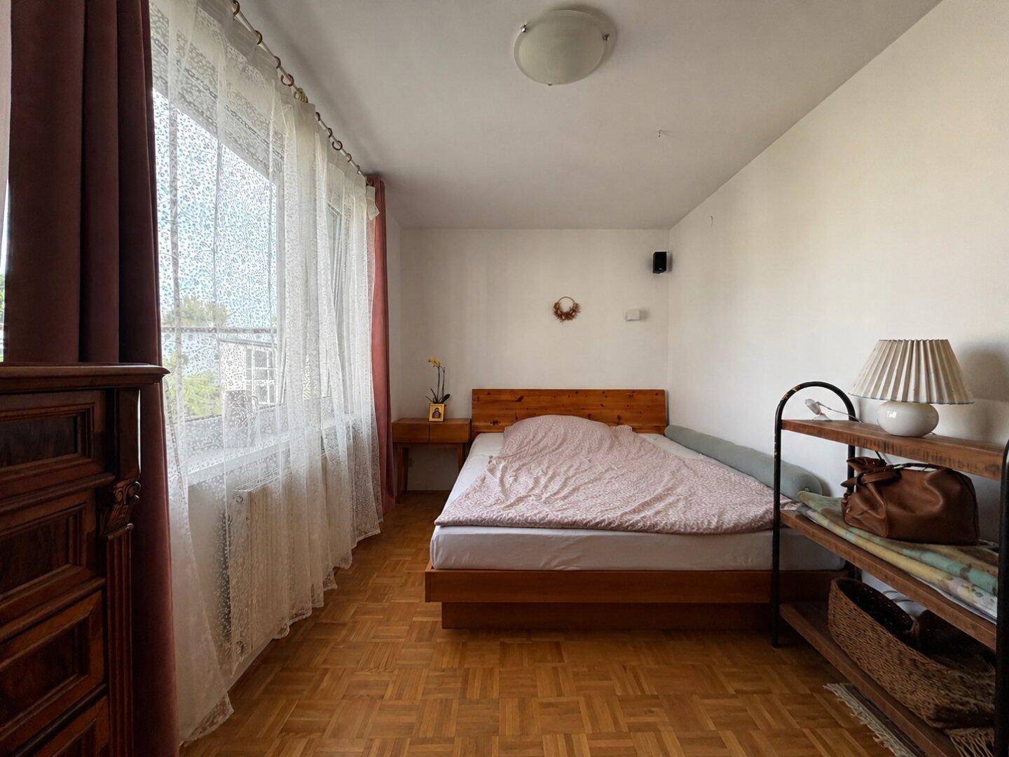 Helles Schlafzimmer mit Holzparkettboden, großem Fenster und einfacher Einrichtung.