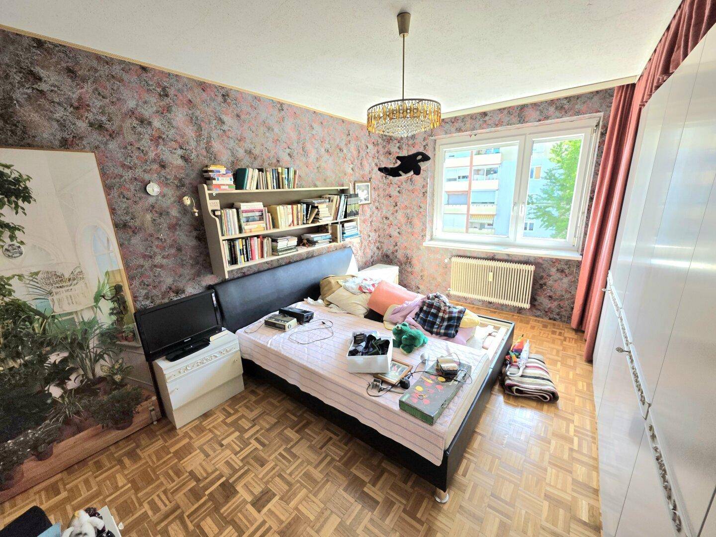 Schlafzimmer mit gemusterter Tapete, Bett, Bücherregal und Blick ins Grüne.