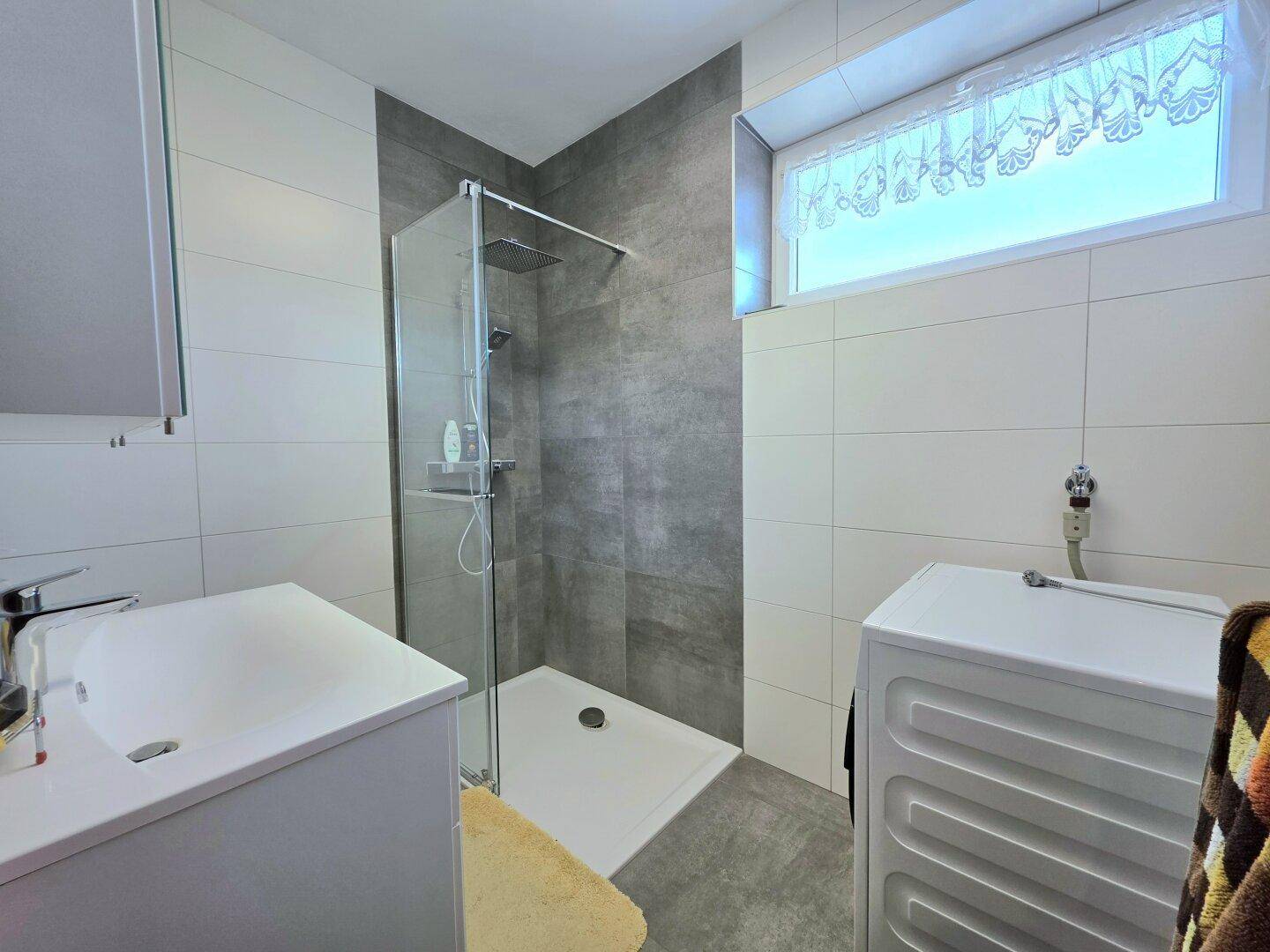 Modernes Badezimmer mit grauen und weißen Fliesen, ebenerdiger Dusche und Fenster.