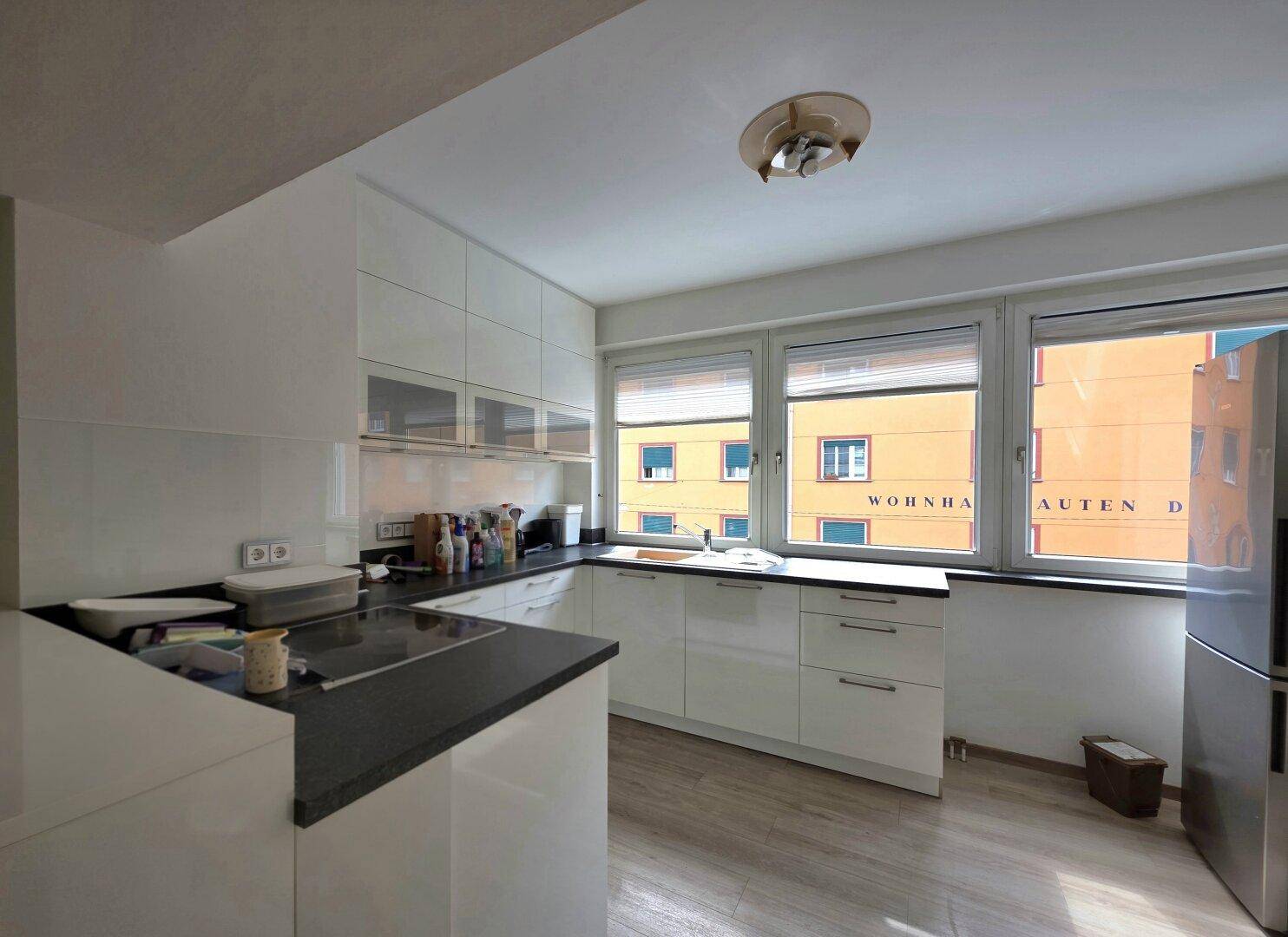 Helle Küche mit weißen Hochglanzfronten, dunkler Arbeitsplatte und Fensterblick auf umliegende Gebäude.