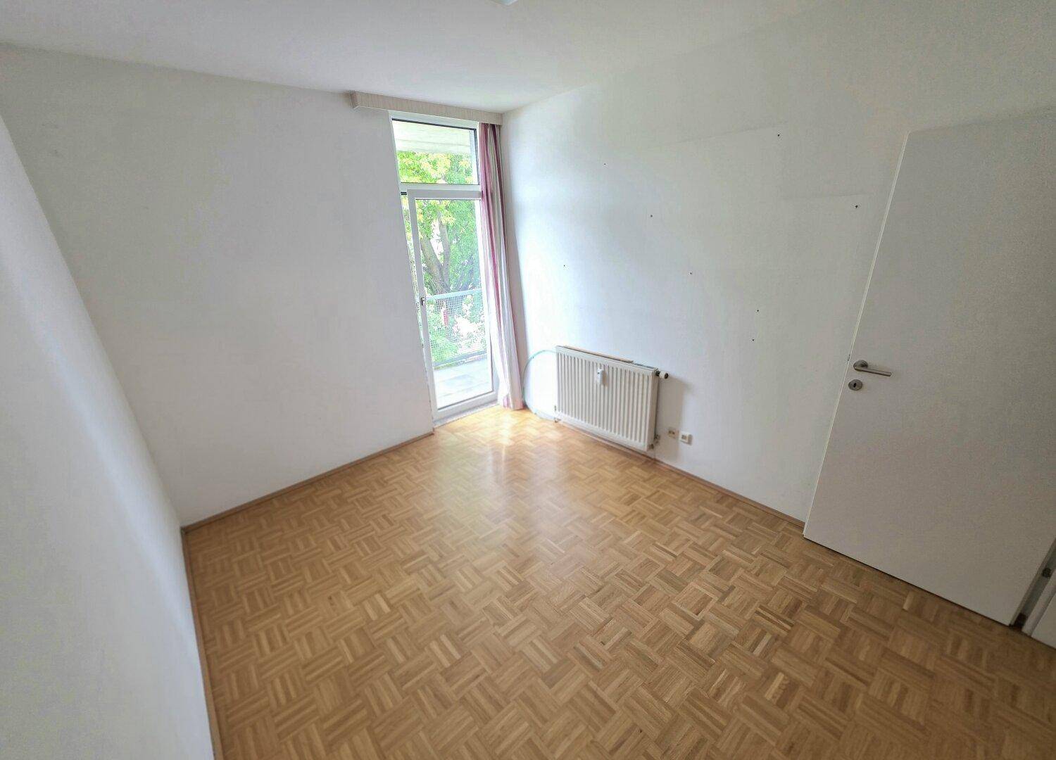 Leeres Schlafzimmer mit Parkettboden und großem Fenster, das viel Tageslicht hereinlässt.