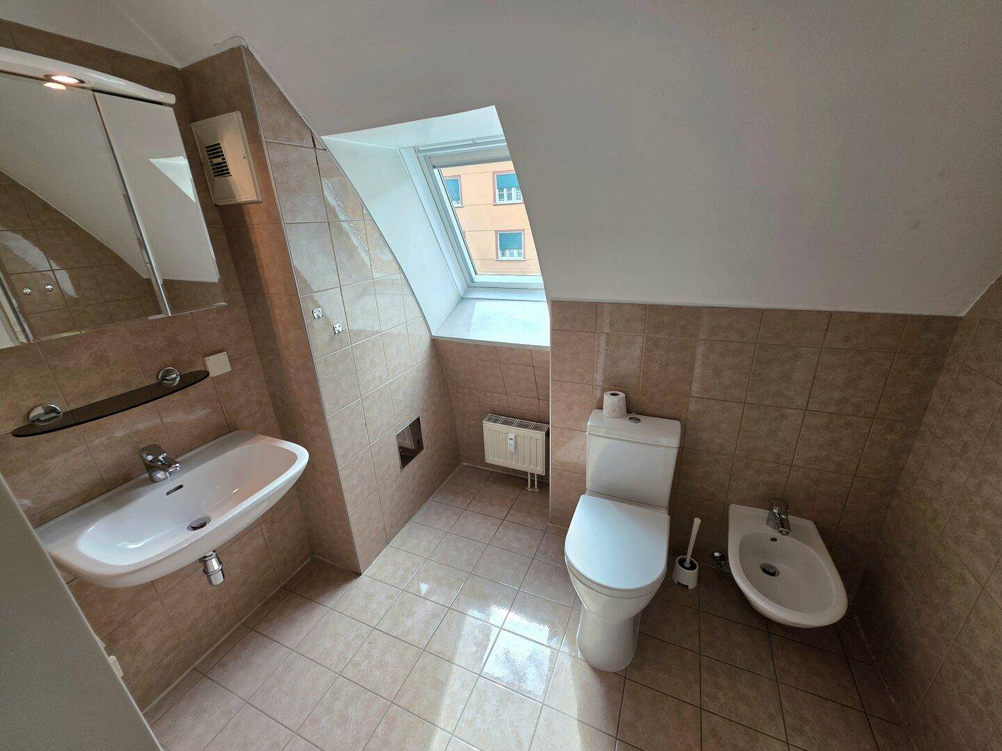 Badezimmer mit Dachfenster, Fliesenboden, Waschbecken, Toilette und Bidet.