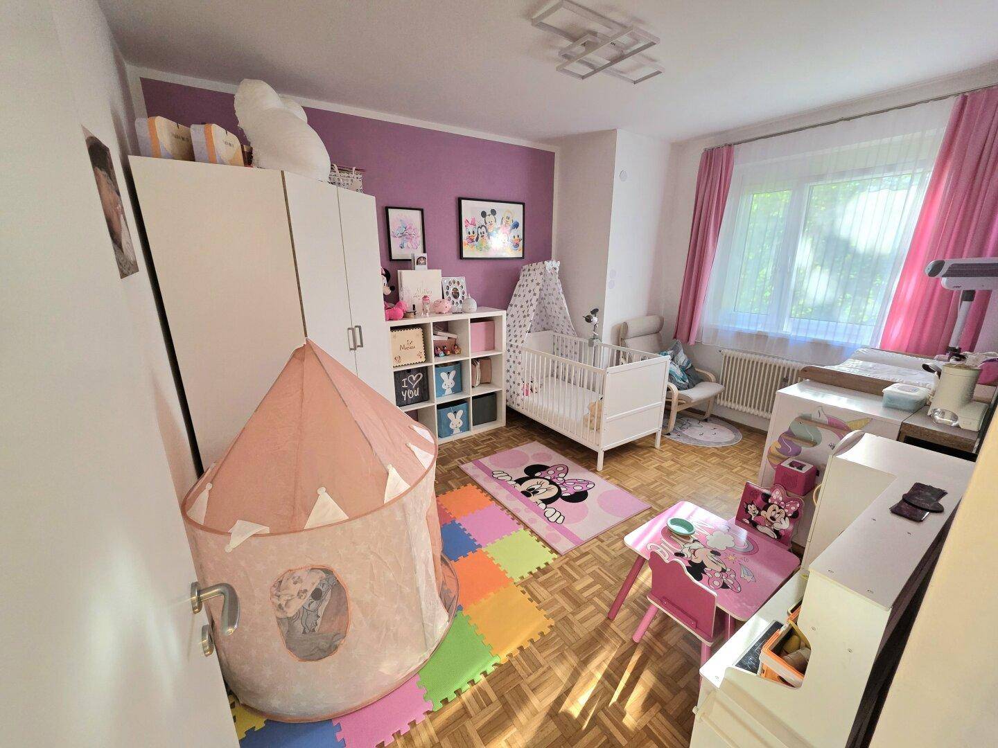 Kinderzimmer mit lila Akzentwand, Babybett, Spielzelt und rosa Vorhängen.