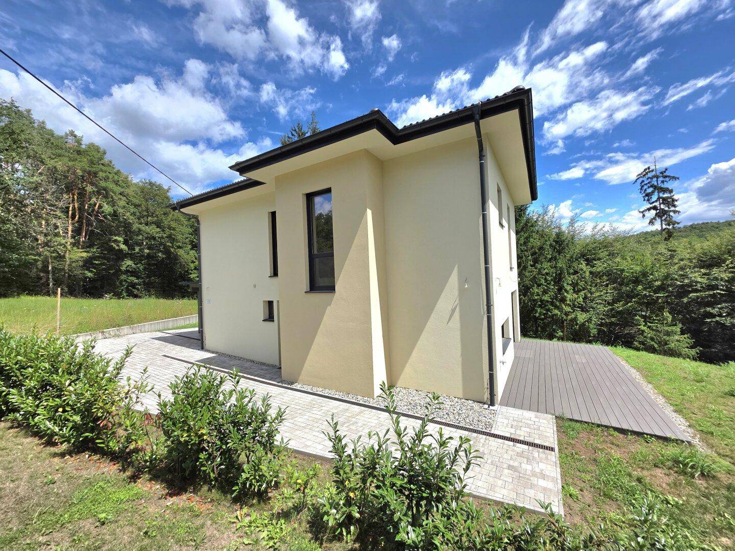 Seitenansicht des modernen Hauses mit gepflastertem Zugang und umgebender Natur unter blauem Himmel.