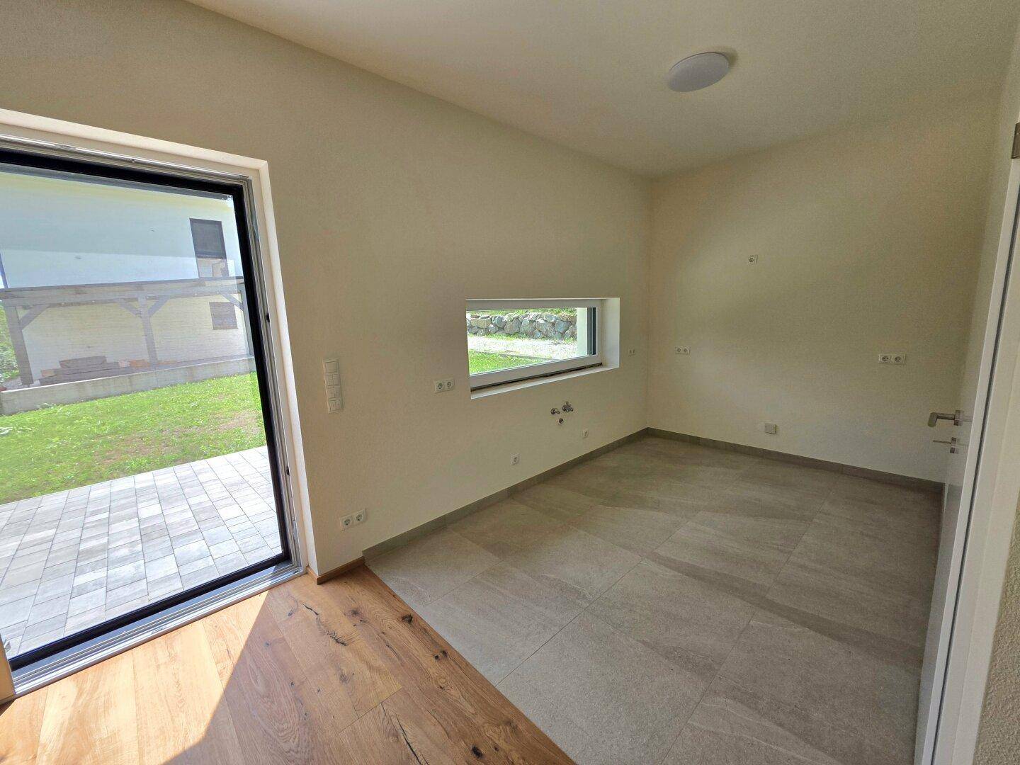 Heller Raum mit Holzboden und Fliesenbereich, großer Fensterfront und Zugang zum Außenbereich.