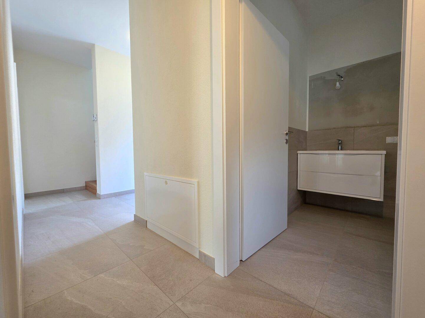 Heller Flur mit Fliesenboden, der zu einem modernen Badezimmer mit Waschtisch führt.
