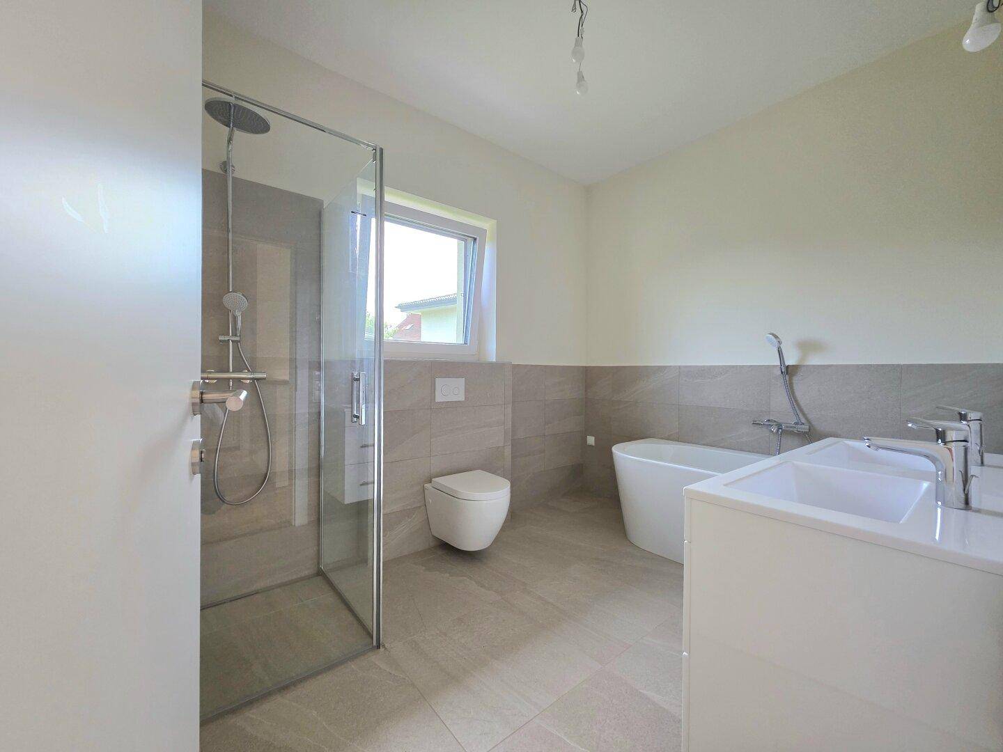 Modernes Badezimmer mit Dusche, Badewanne, Doppelwaschbecken und Fenster, hell gefliest.