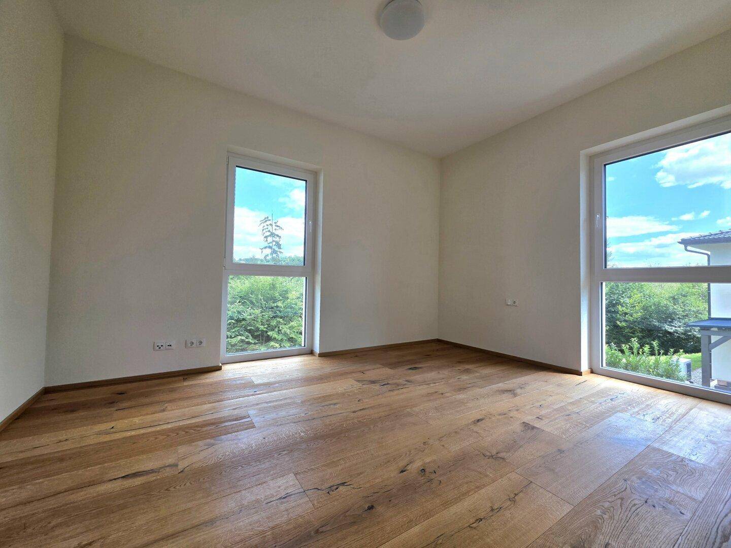Helles Schlafzimmer mit Holzboden und zwei Fenstern, die einen Blick ins Grüne bieten.