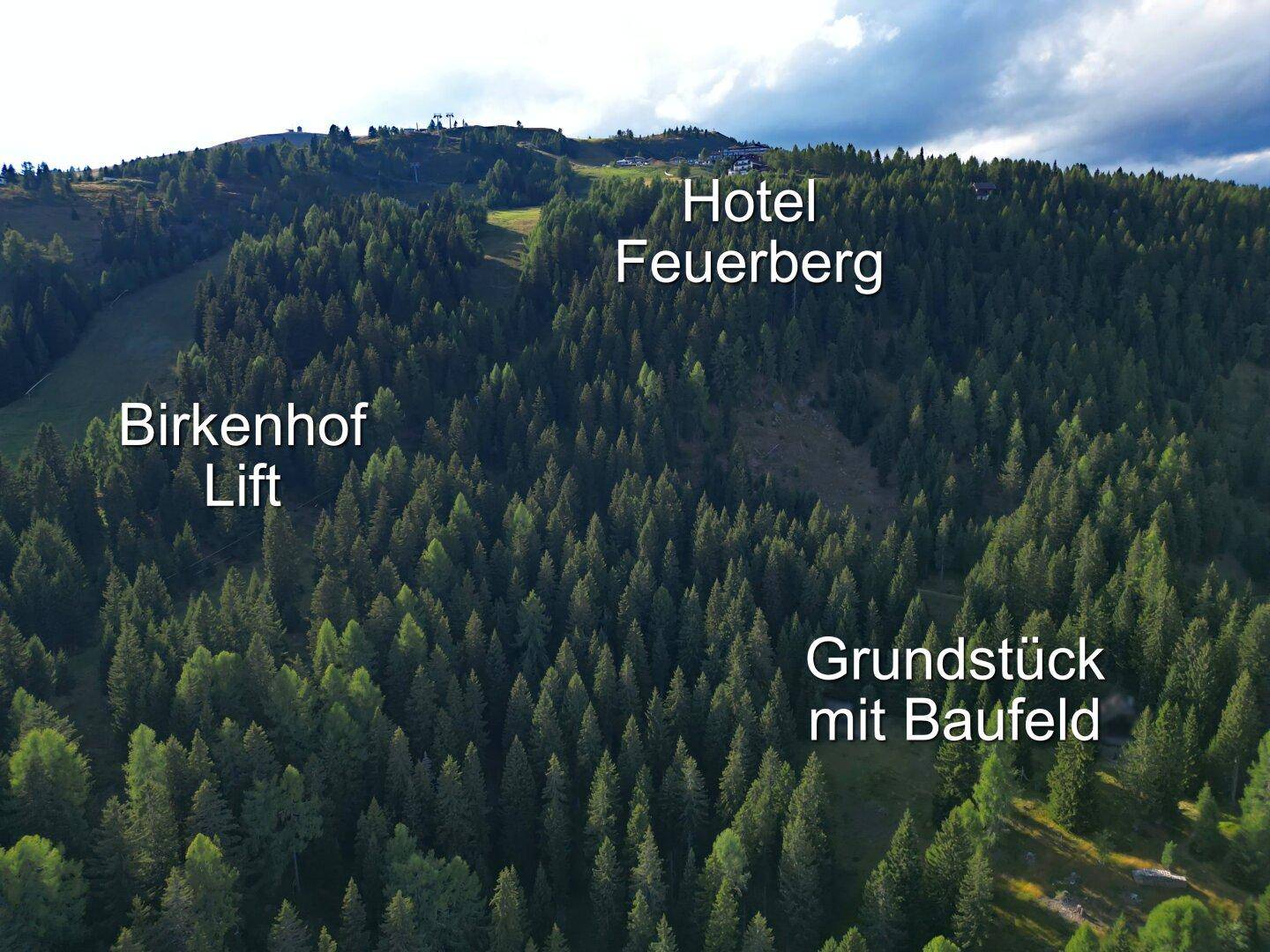 Luftaufnahme eines bewaldeten Berghangs mit Beschriftungen, die auf ein Hotel, einen Lift und ein Baufeld hinweisen.