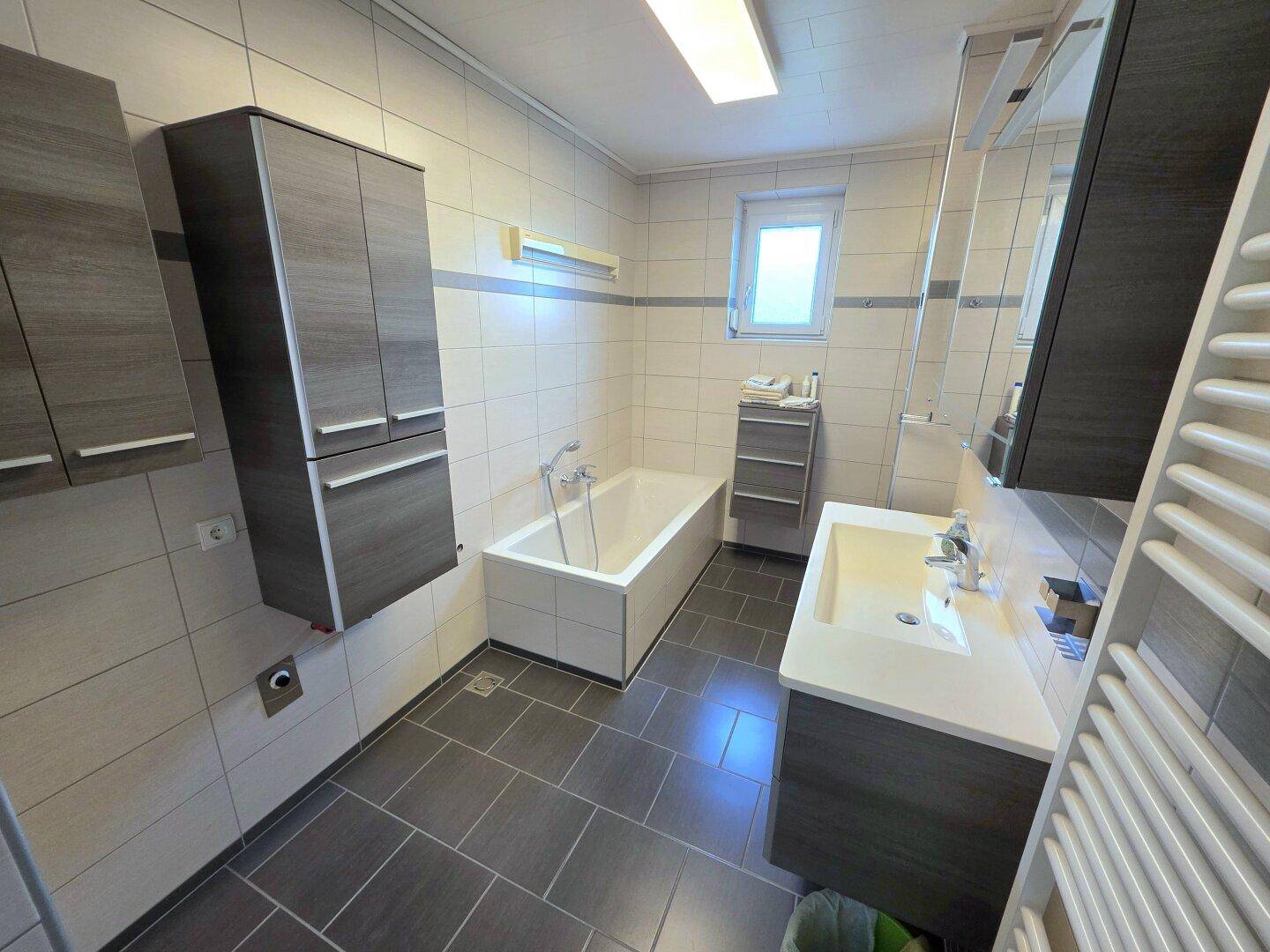 Modernes Badezimmer mit Badewanne, Waschtisch und grauen Fliesen, inklusive Fenster.