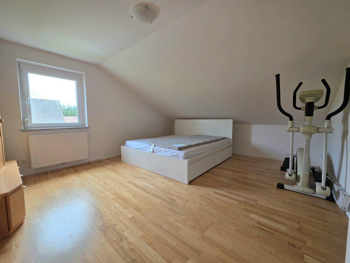 Schlafzimmer im Dachgeschoss mit Holzboden, Bett und Fitnessgerät unter der Dachschräge.